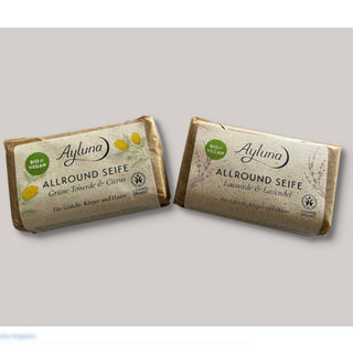 Two bars of Ayluna Allround Seife soap wrapped in kraft paper, one with yellow lemon illustrations and the other with lavender illustrations, on a light gray background.