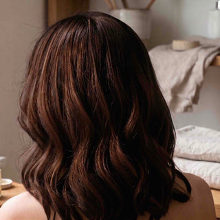 A close-up back view of a person with shoulder-length, wavy brown hair with subtle warm highlights, set against a softly blurred indoor background with neutral-toned towels and furniture.