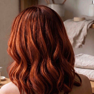 Wavy medium-length auburn-red hair shown from the back on a person in a softly lit indoor setting.
