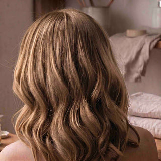 A close-up back view of a person with shoulder-length, wavy hair in a warm light brown or caramel shade, set against a softly blurred indoor background.