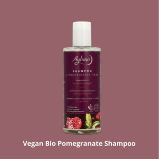 A bottle of Ayluna Vegan Bio Pomegranate Shampoo centered on a solid dusty rose background, with white text below reading 'Vegan Bio Pomegranate Shampoo'. The bottle has a white cap and a dark purple label with pomegranate and green leaf illustrations.