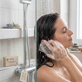 A woman with wet dark hair washes her hair in a shower while wearing a gray sponge. A box of Aylvong Almond Soap is visible on the shelf behind her.