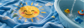 A close-up of a blue plush towel with a large smiling yellow sun and small white clouds, next to a light blue plastic tub with water running in and bath toys inside.