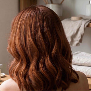 A person shown from the back with shoulder-length wavy hair in a warm coppery auburn tone, photographed indoors with softly blurred neutral-toned background elements.