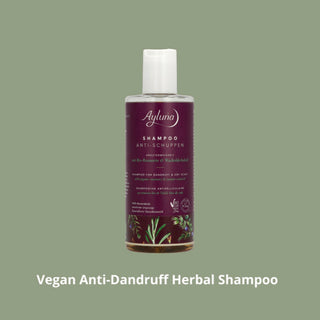 A bottle of Ayluno Vegan Anti-Dandruff Herbal Shampoo centered on a muted sage green background, with white text below reading 'Vegan Anti-Dandruff Herbal Shampoo'. The product has a white cap and a deep purple label with botanical illustrations near the bottom.