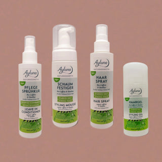 Four Ayluna hair care products arranged on a dusty pink background: a leave-in conditioner spray bottle, a styling mousse bottle, a hair spray bottle, and a styling gel stick.