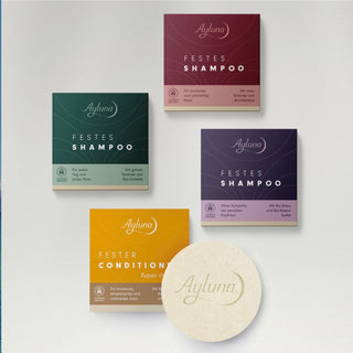 A set of Ayluna haircare products arranged on a light gray background: two boxed shampoos in dark green and dark purple, one boxed conditioner in mustard yellow, and one round cream-colored bar labeled Ayluna. No people or real environment are present.